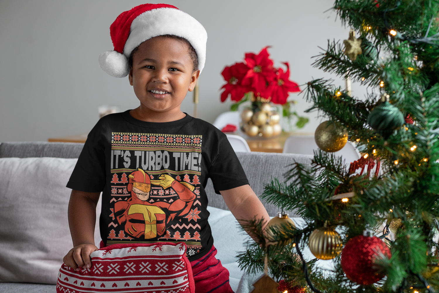 Child wearing a Christmas-themed shirt and Santa hat in front of a decorated Christmas tree.